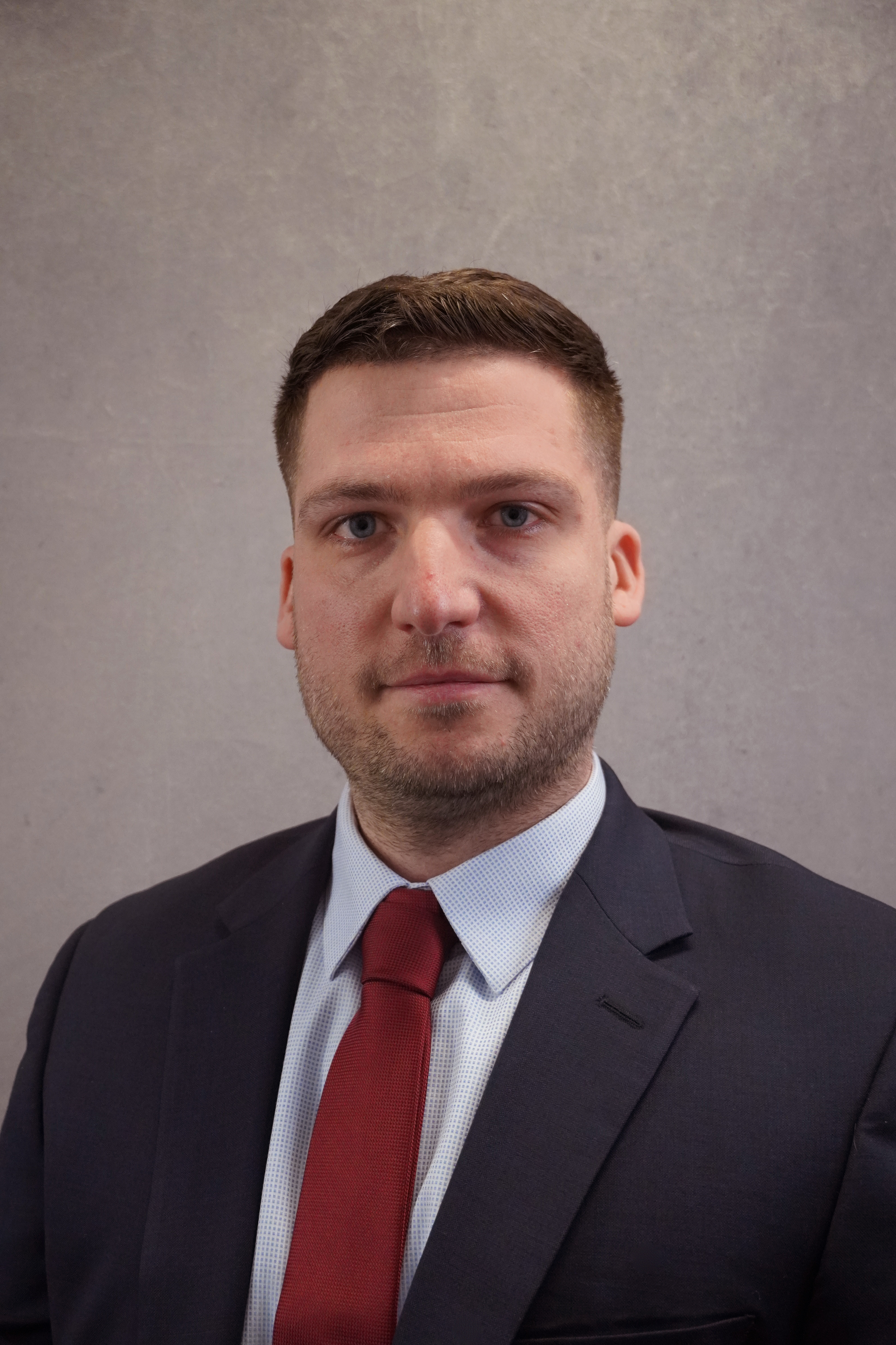 An individual with short brown hair wearing a white button up with a red tie and black suit coat. 