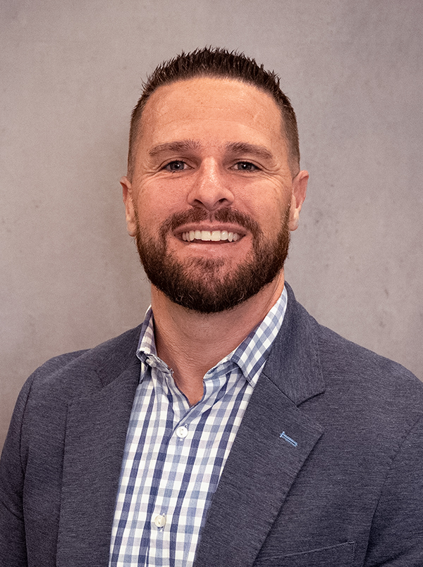 An individual with short brown hair wearing a button up shirt with a grey suit coat.